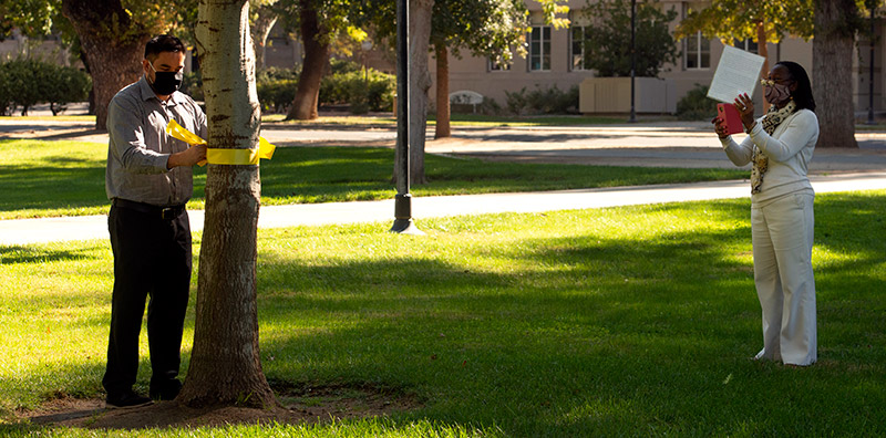 Man tying yellow ribbon on tree as woman films with phone.