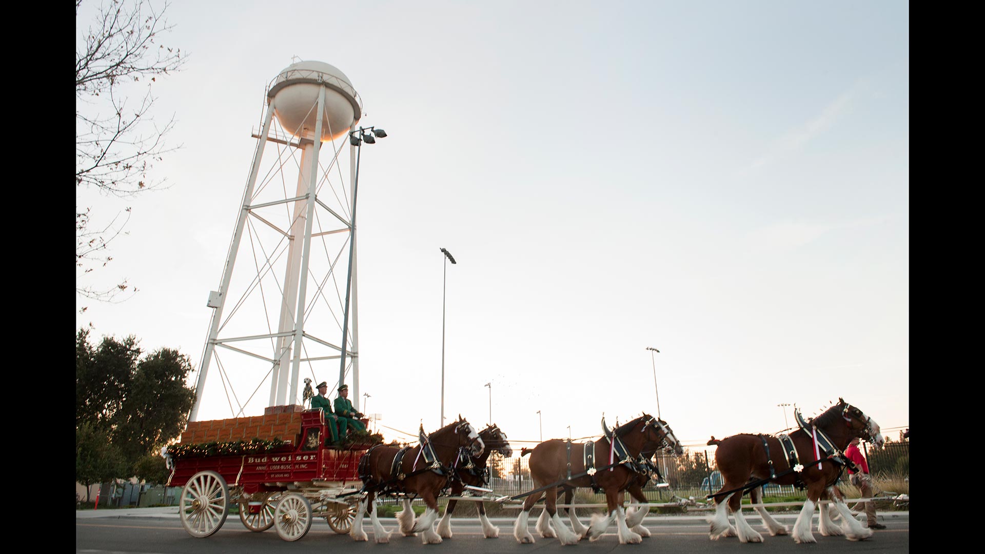 A team of draft horses pulls a large red wagon past one of the UC Davis water towers. Two men in green outfits drive the wagon.