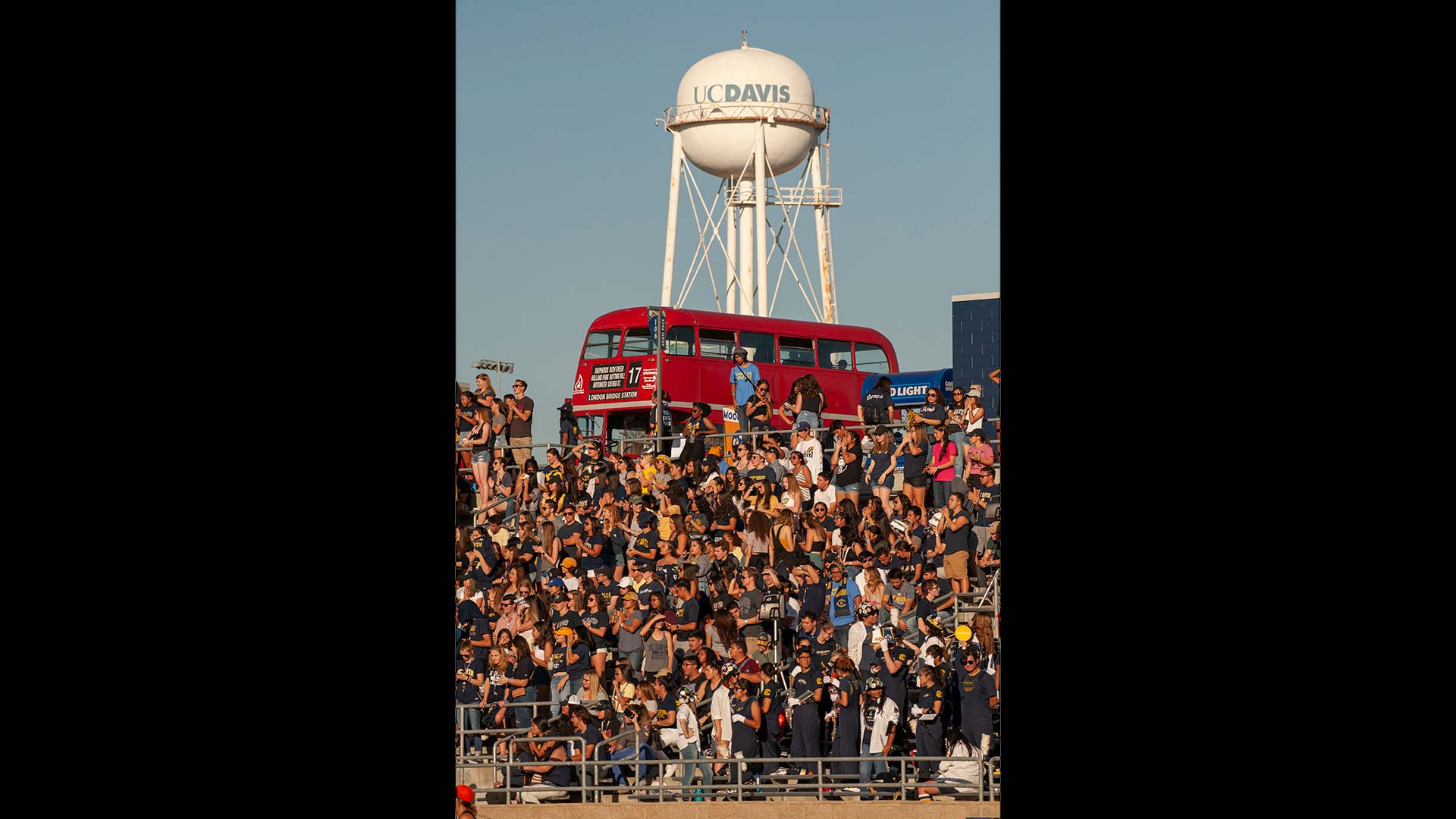 The UC Davis water tower appears behind a full stadium of fans and a Unitrans london bus.