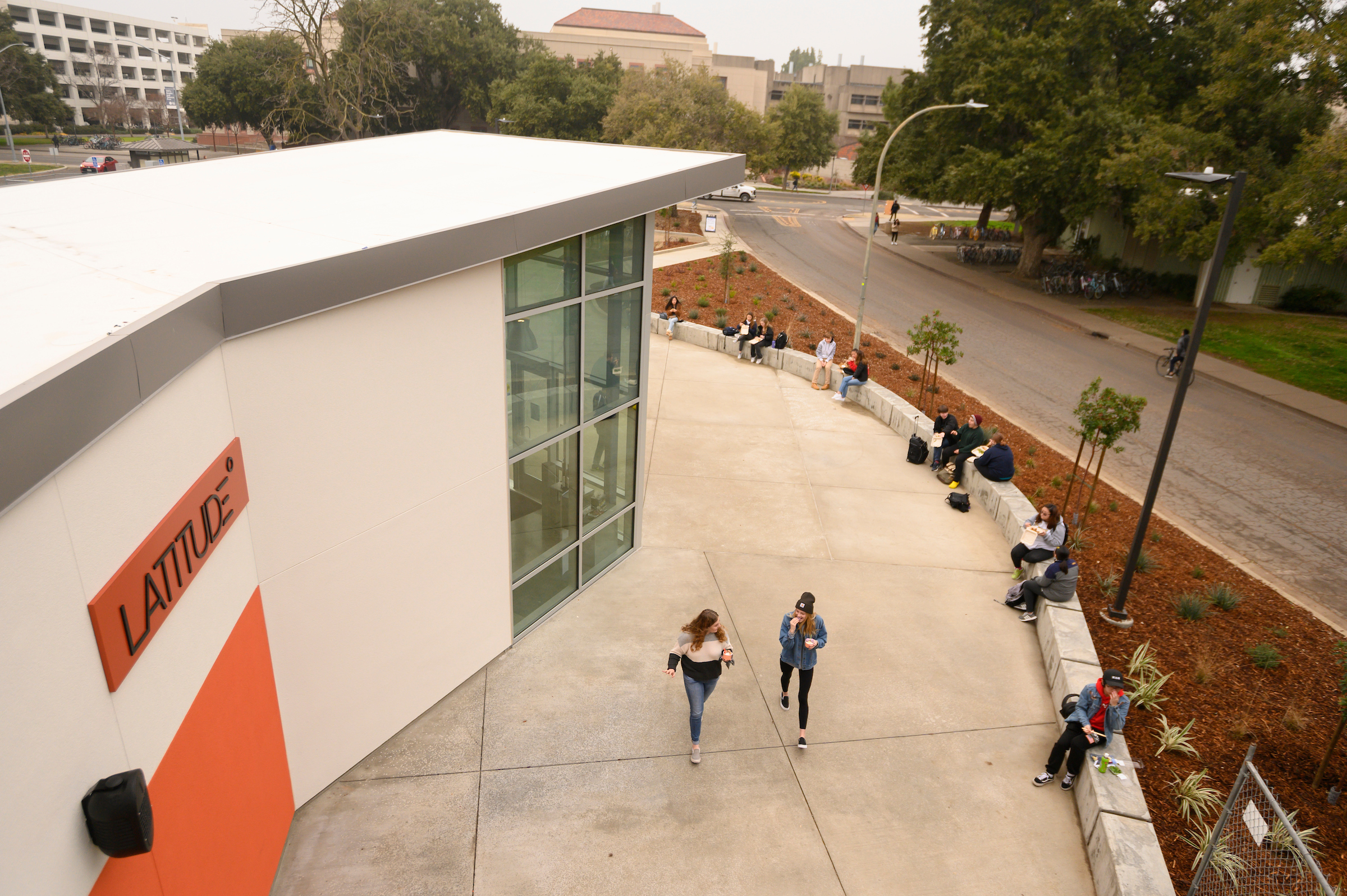 An aerial shot of a white dining commons building with a sign bearing its name, Latitude, as student walk below on the sidwalk.