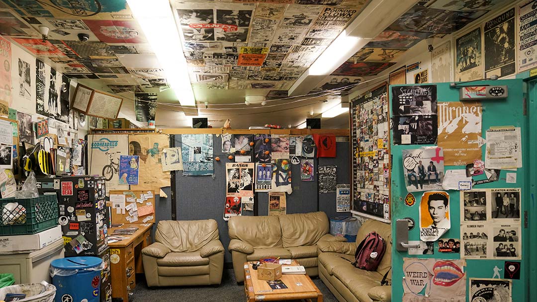 Photo of a cluttered lounge area. The room has two leather couches, a coffee table and a desk, but is also filled with crates and boxes. The walls and ceiling are covered with music flyers and posters.