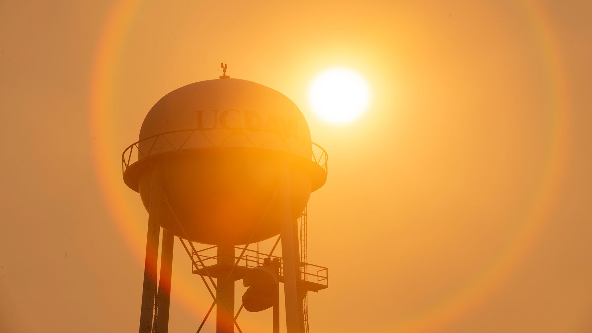 Silhouette of a UC Davis water tower against the sun and an orange sky. A red lens flare surrounds the tower.