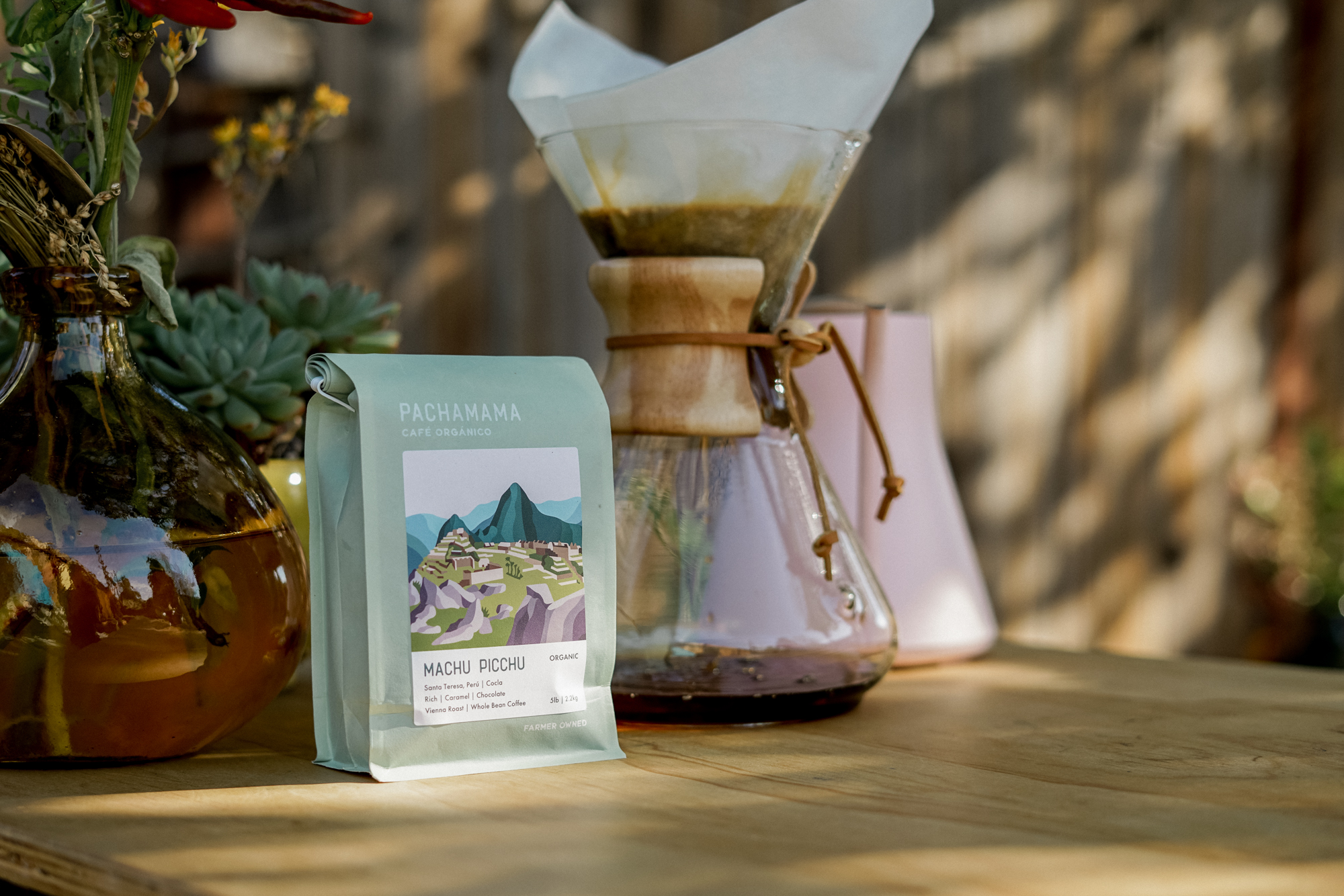 A photo of a bag of Pachama coffee grounds sits next to a pour over carafe of coffee and a pink electric kettle behind it. 