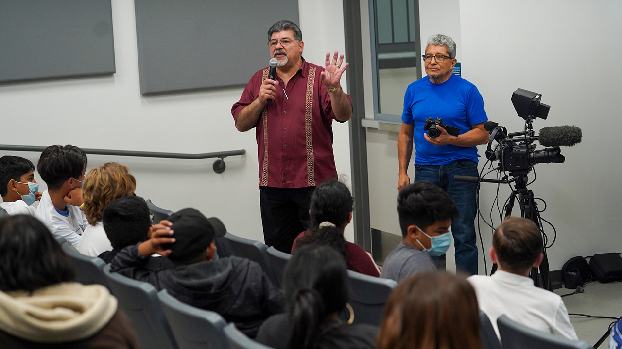 Two men with video equipment talking to seated students