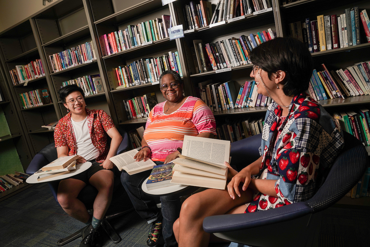 Two students and a library laugh with open books in front of a library shelf at UC Davis.