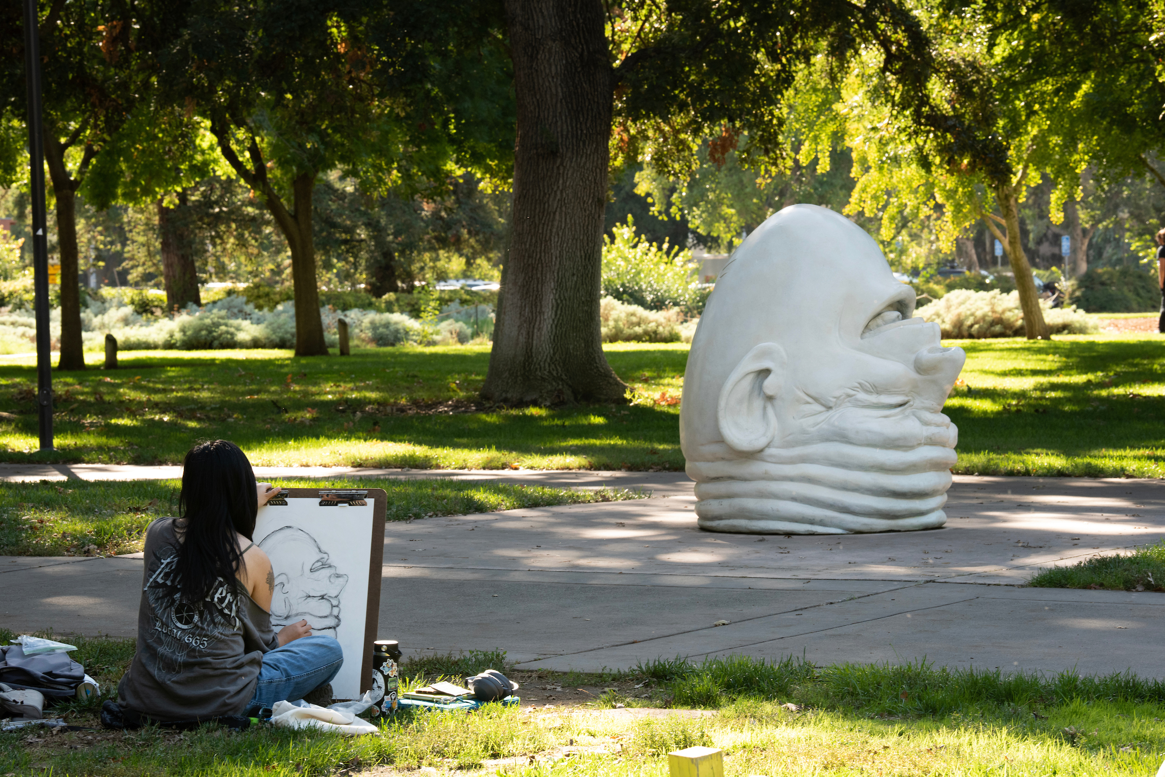 A student sits outside Mrak Hall on a sunny day sketching a large drawing of the “Egghead sculpture Eye on Mrak (Fatal Laff)."