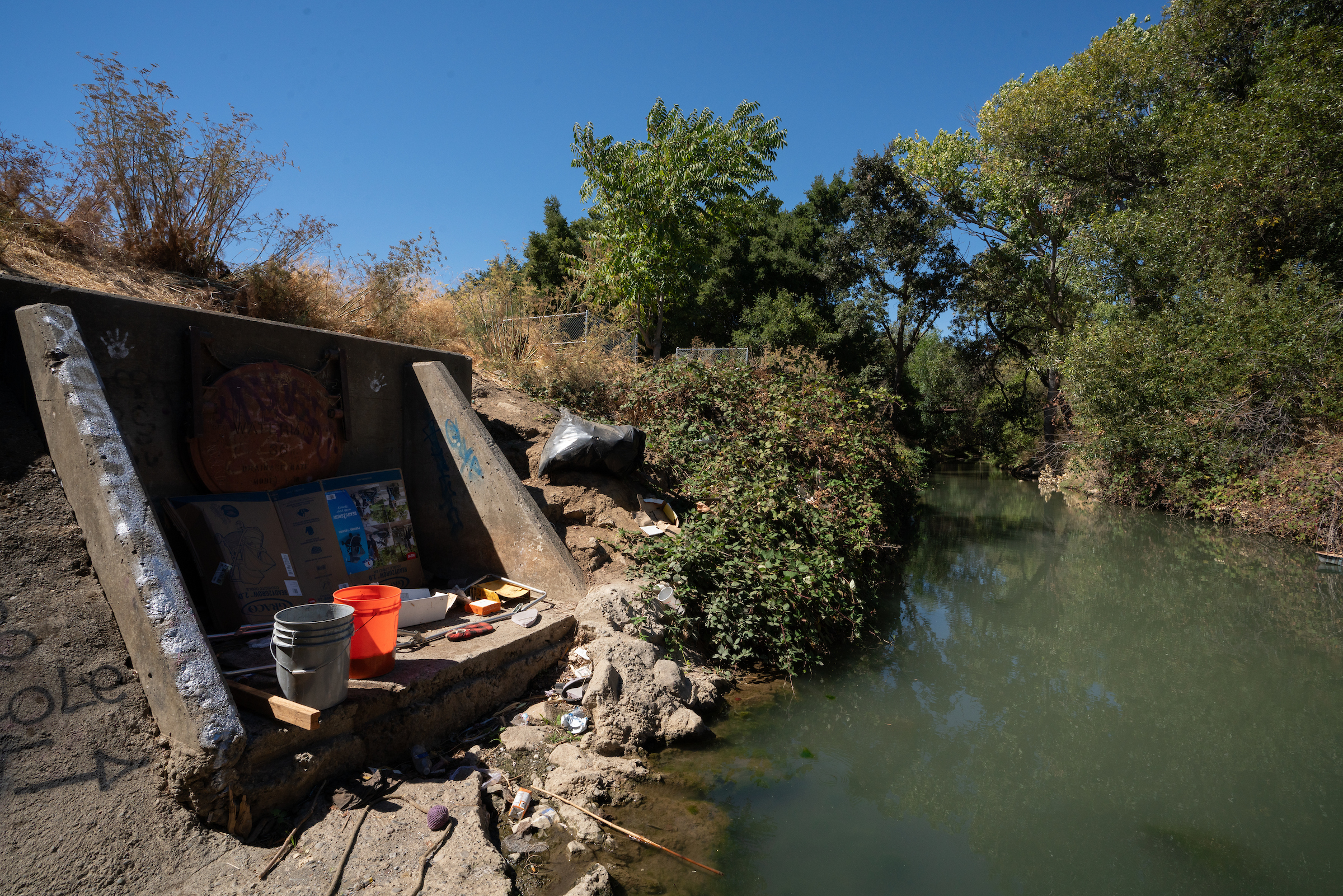 A green, tree-lined creek meets a tunnel underpass holding a cardboard box, two large buckets and other objects