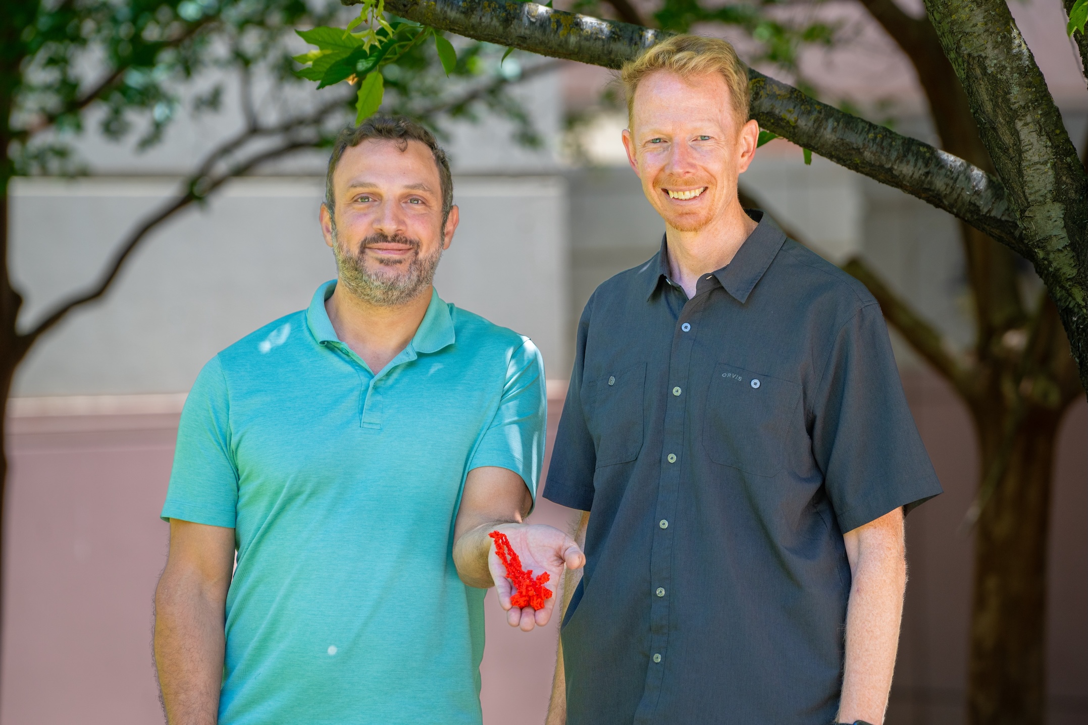 Two smiling men standing under a tree, one holding a glass of red drink.