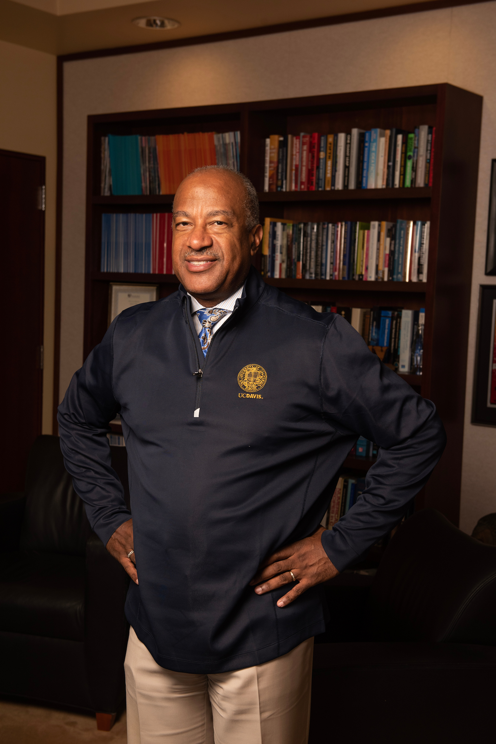 Smiling older man standing with hands on hips in office, wearing a navy pullover bearing the UC Davis seal, standing in front of a large bookshelf