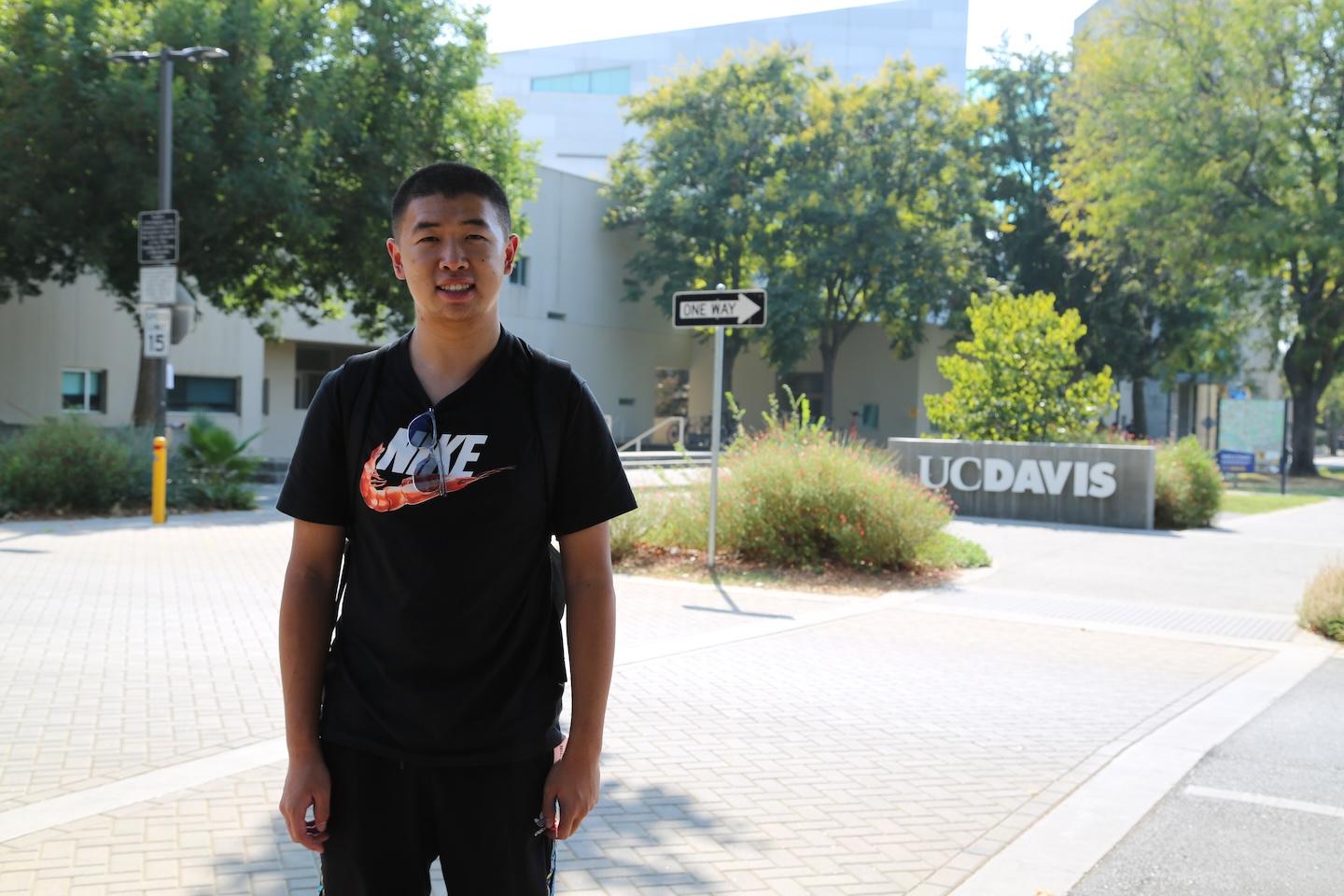 A student stands and smiles near the UC Davis sign, in a black Nike shirt and backpack. Trees, campus buildings and a one-way street sign are visible in the background.