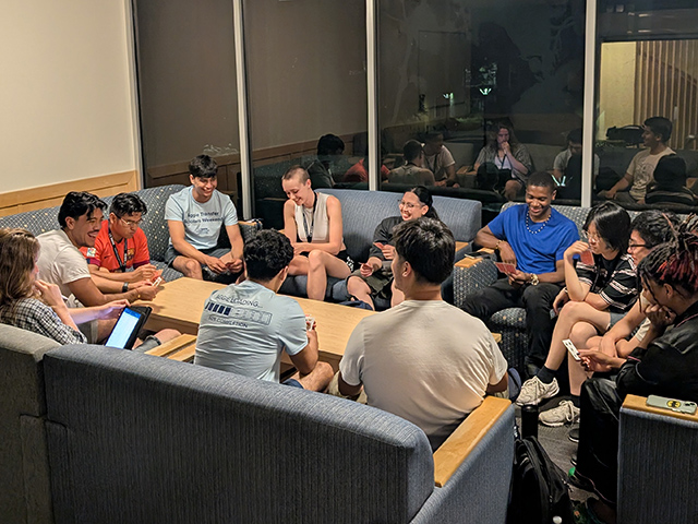About a dozen young adults seated in a living room around a coffee table, engaged in discussion.