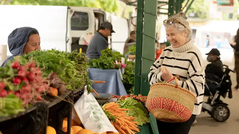 Woman in striped sweater with woven basket selecting carrots at outdoor farmers market