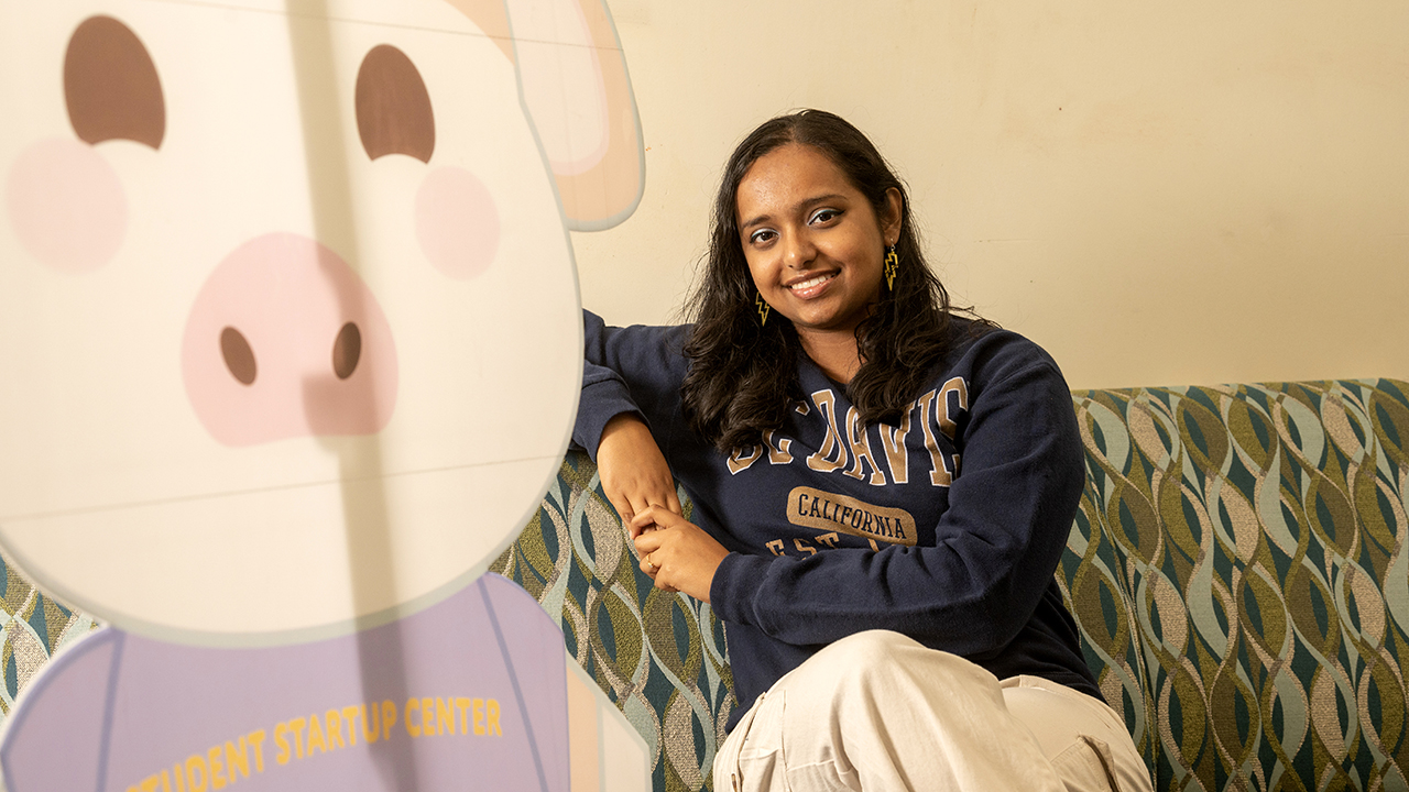 Young woman in navy UC Davis sweatshirt sitting on a sofa, beside a large cardboard cut-out of the Student Startup Center's mascot.