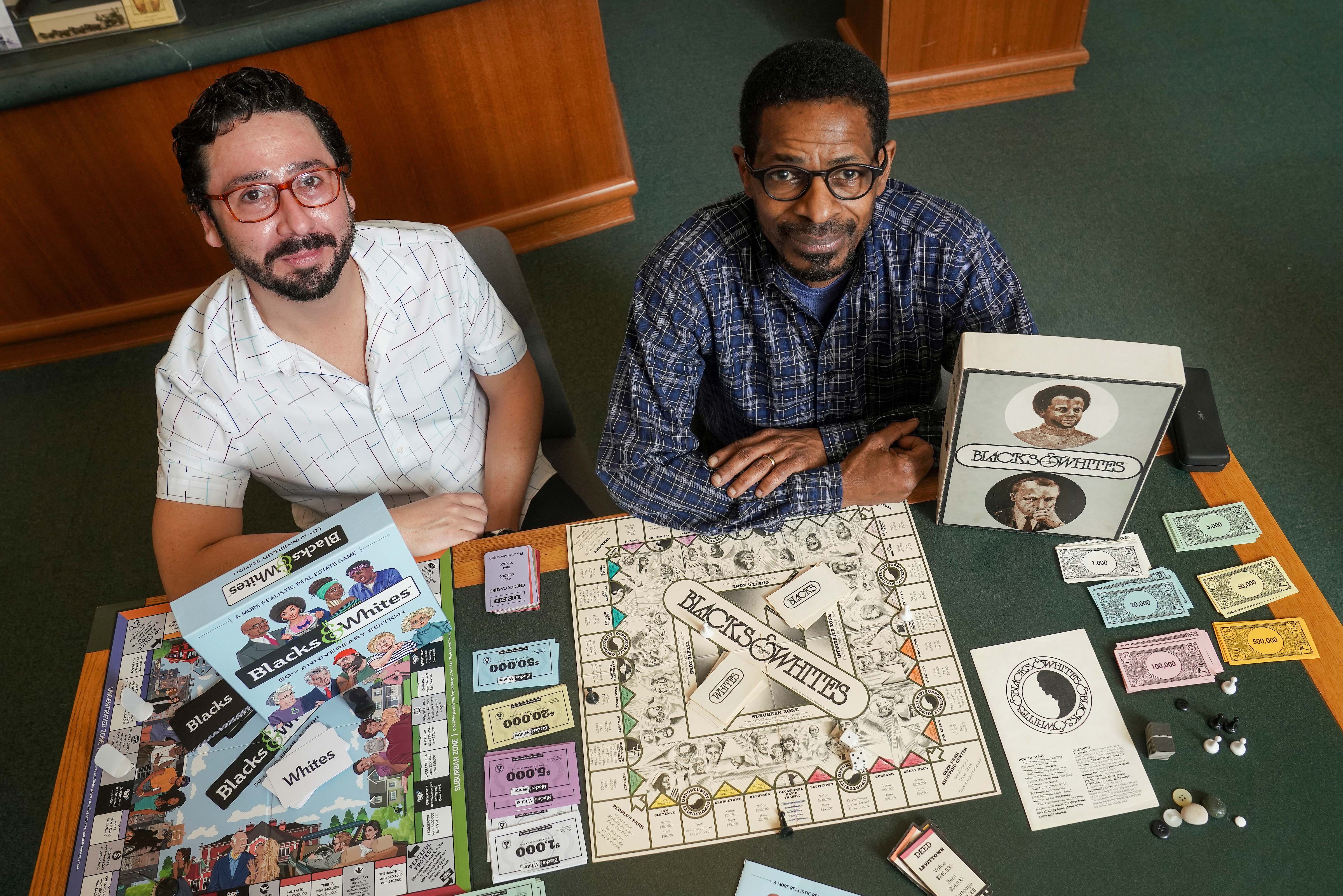 Two people at a table with board games on it look up at a camera w