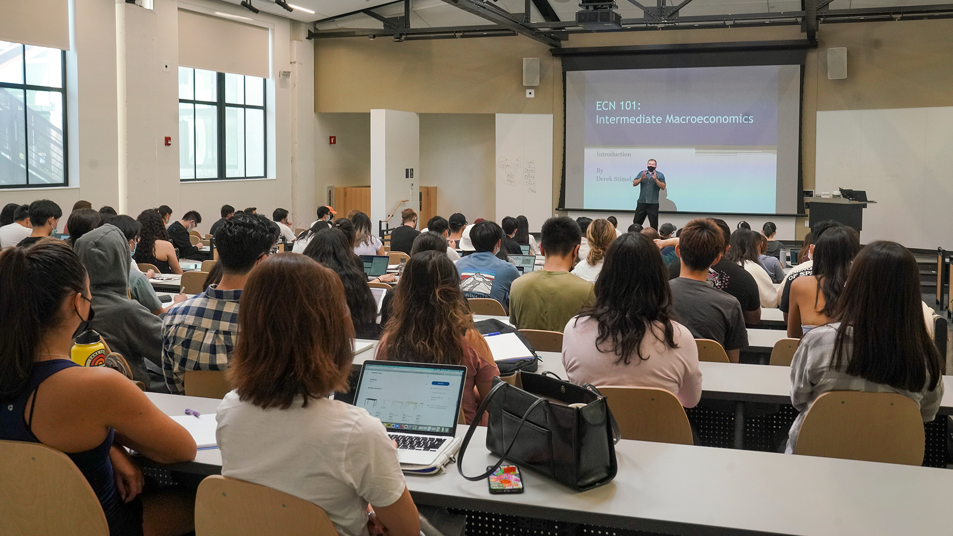 Photo inside a classroom during class. The camera is at the back of the room, showing the backs of students as they listen to a lecturer at the front. There is a large screen pulled down from the ceiling across the front and large windows on one side of the room.