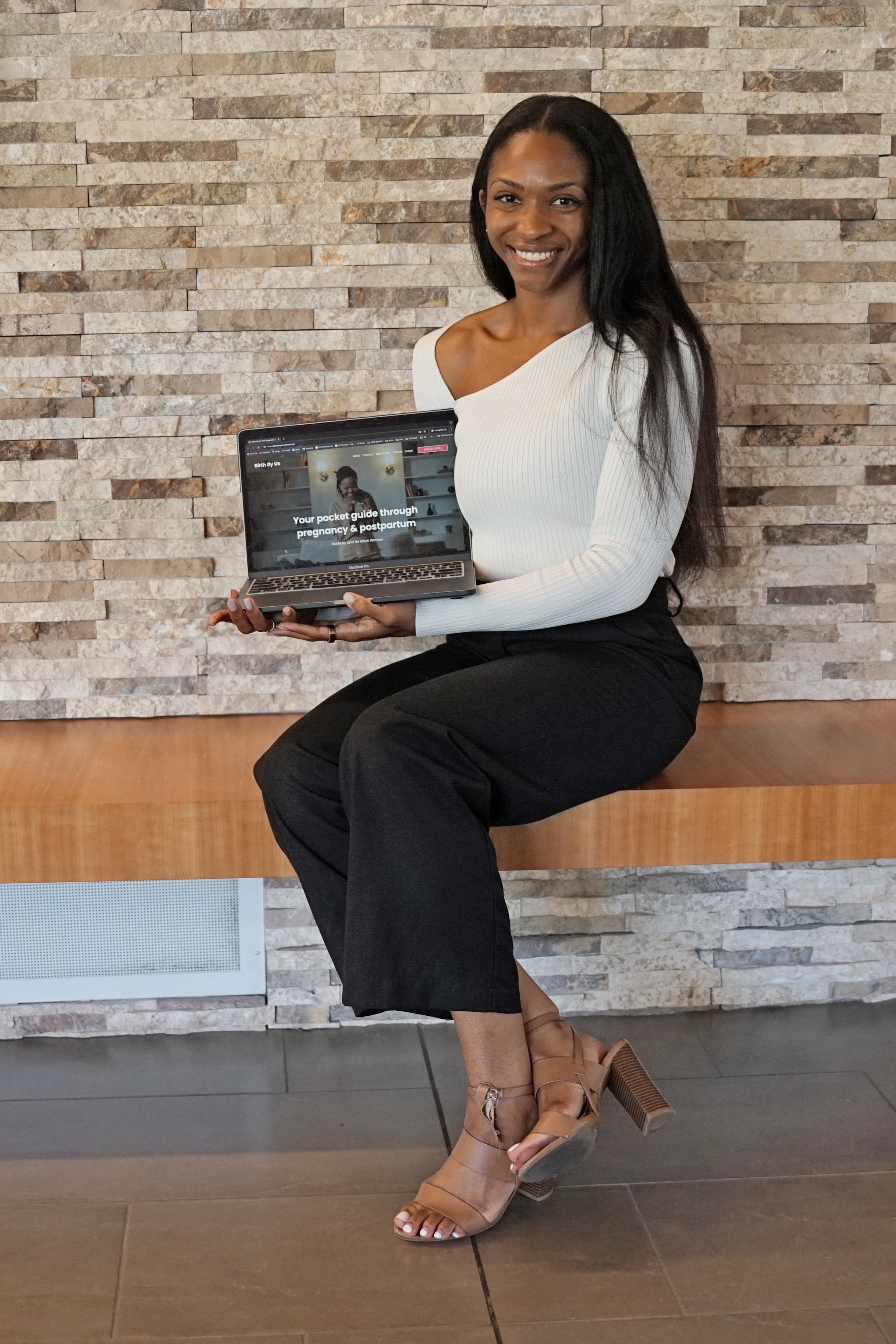Black woman holds laptop illustrating software. She is pictured in front of a wall wearing a white blouse and black pants
