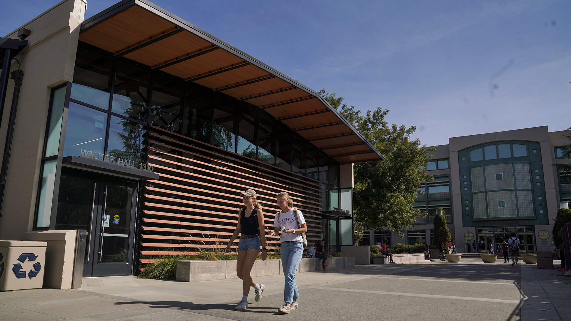 Photo showing a building facade at an angle and from a low vantage point. The building has windows on the side edges and top, with large wooden eaves and wood slats on the walls. Two female students walk by toward the camera. Shields Library can be seen in the background.