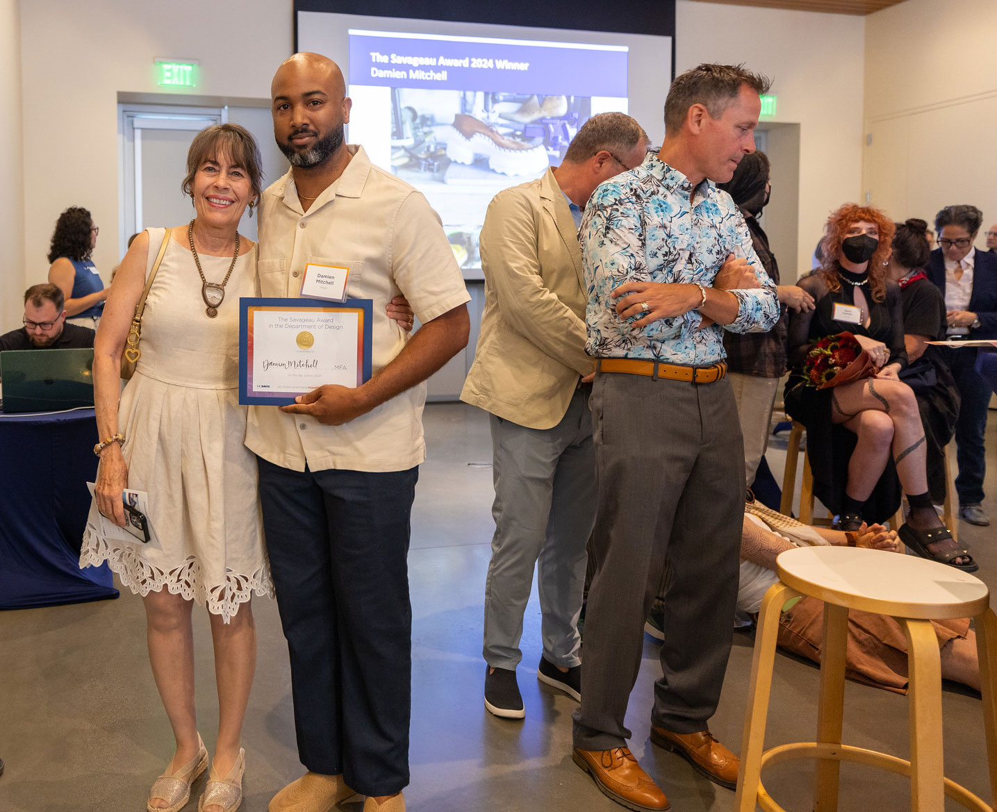 Woman in dress stands next to dark-complected man with award in hand with museum crowd in background