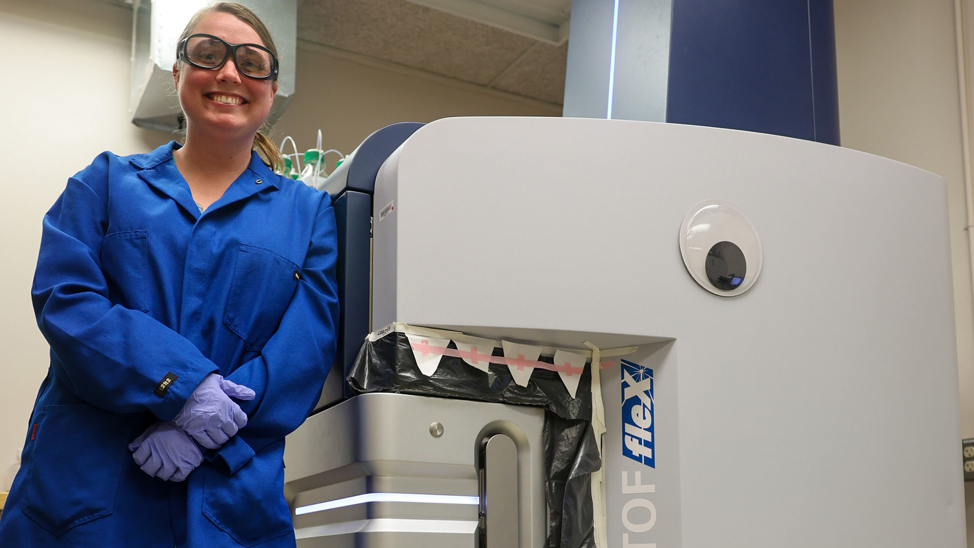 Smiling lab worker in blue coat beside machine decorated with googly eye and paper teeth, photo