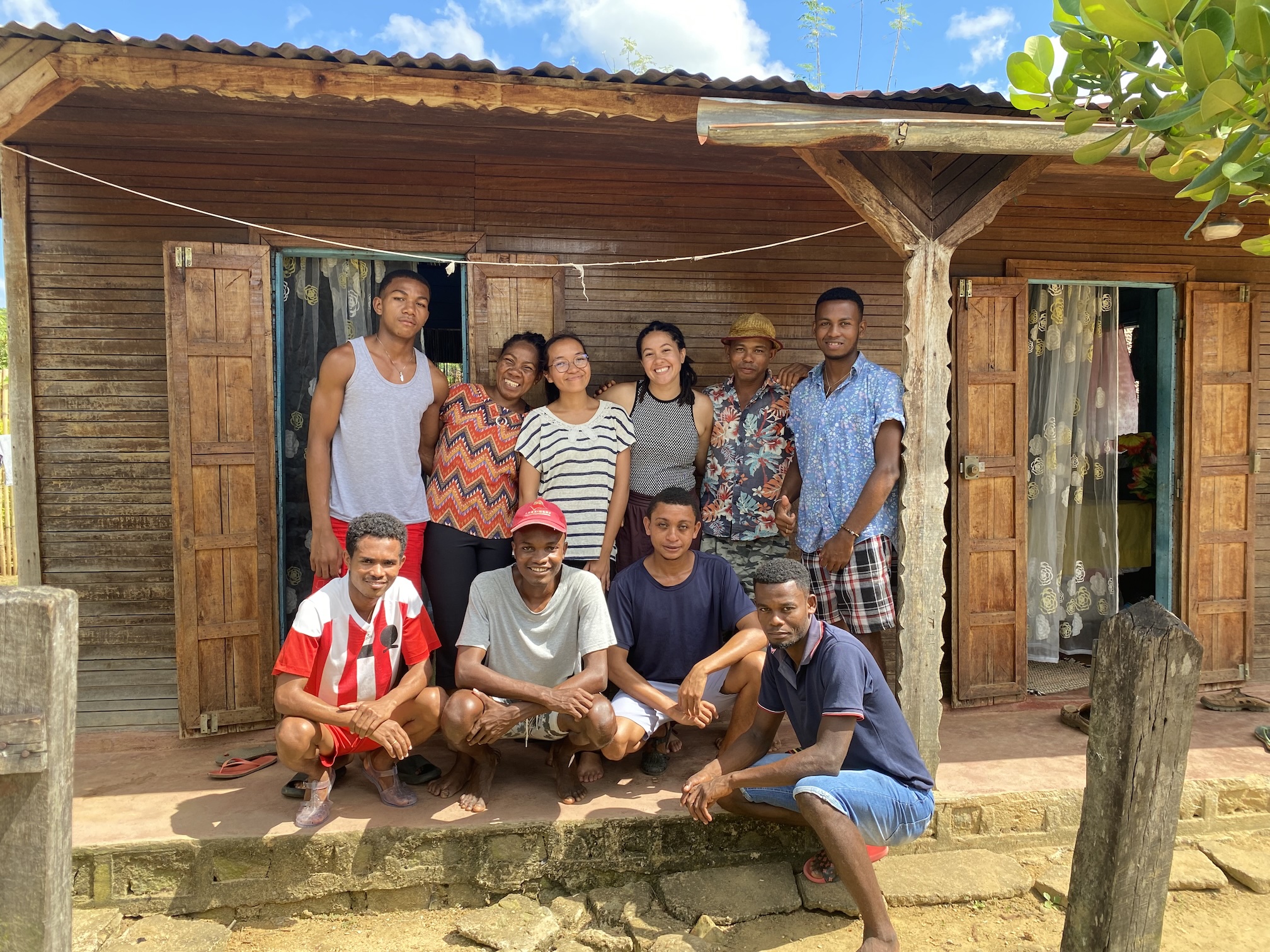 Ten people smile under a wooden balcony and house in Madagascar