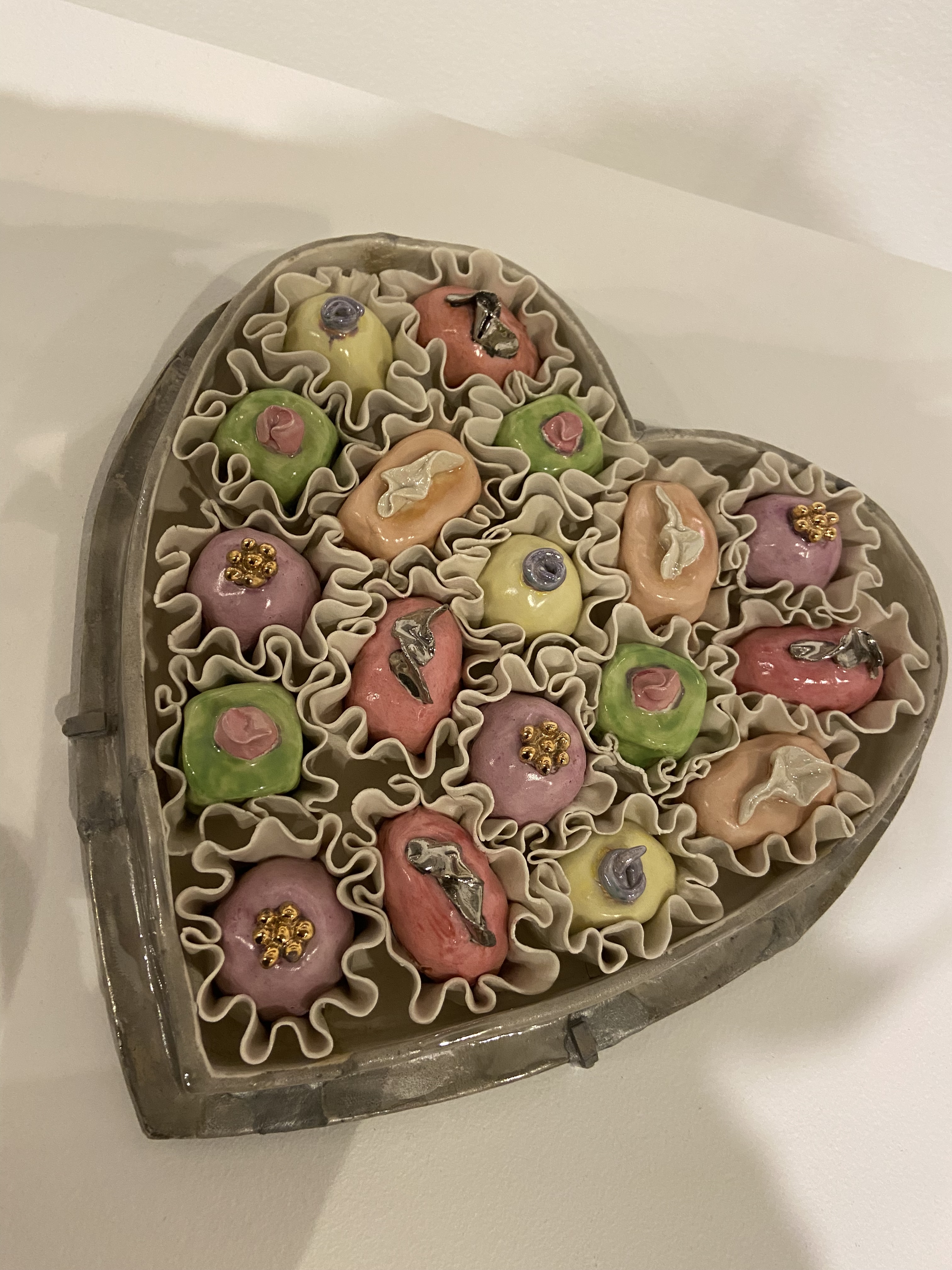 Ceramic depiction of heart-shaped box filled with colorful, decorative floral-shaped confections.