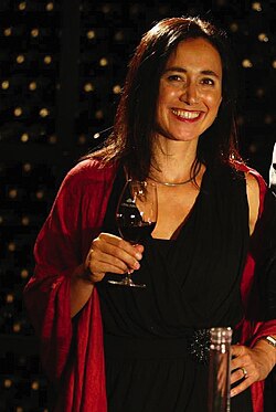 Portrait of a woman standing with a glass of wine, and appears to be in front of a rack of wine bottles