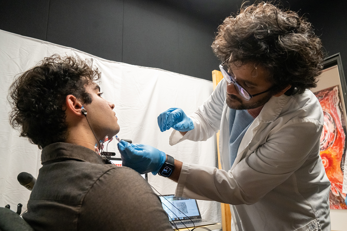 Left, profile of a seated young man with dark curly hair. To the right is a young man with dark curly hair white lab coat and blue gloves putting an electrode on the seated man's jaw.