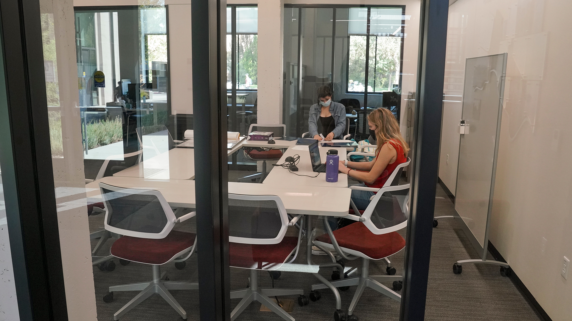 Photo looking through interior glass doors into a room with tables, chairs and two women seated while working on laptops.