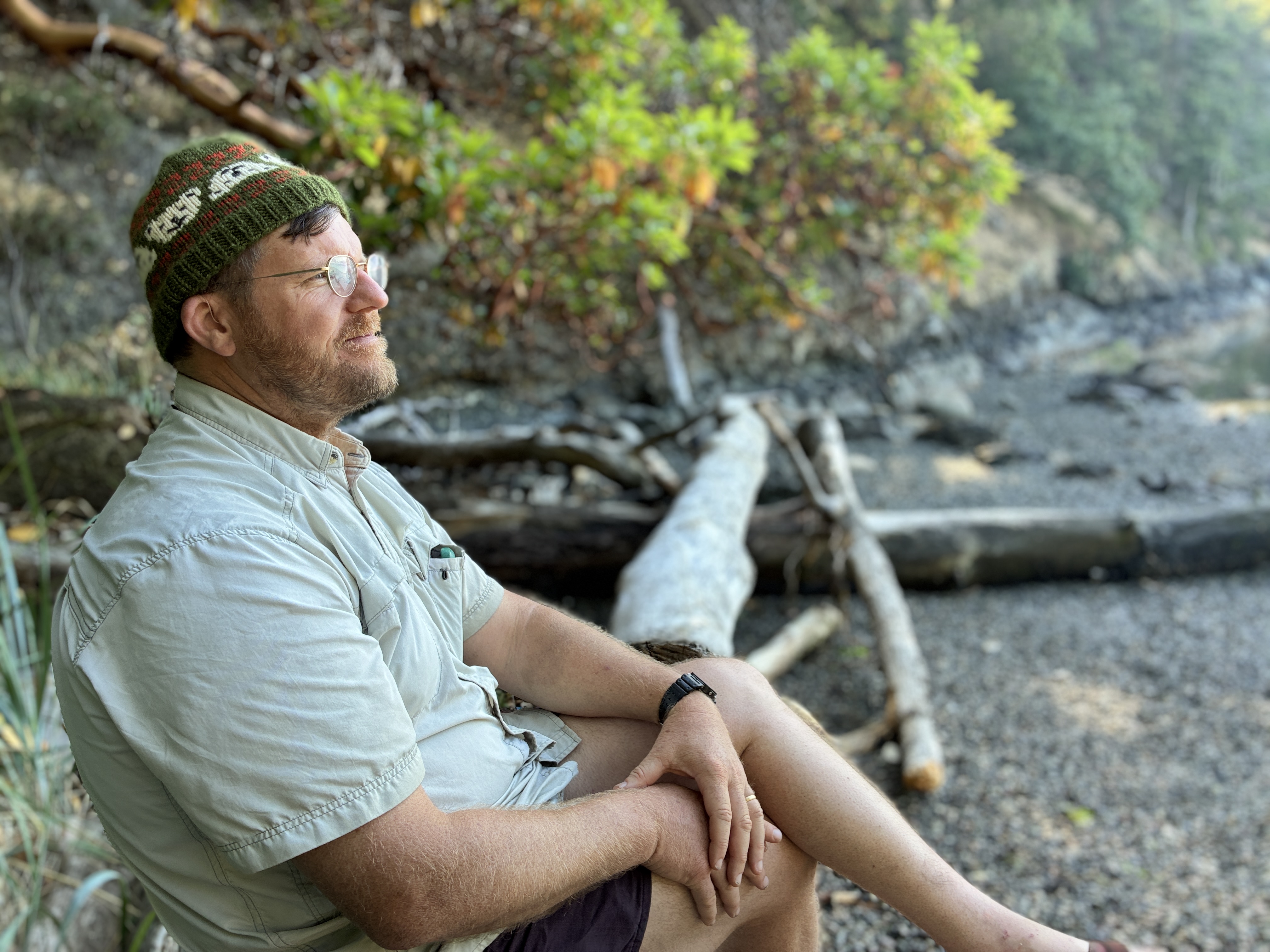 Bearded man in green beanie sitting on driftwood along a rocky, tree-lined shore, pensive