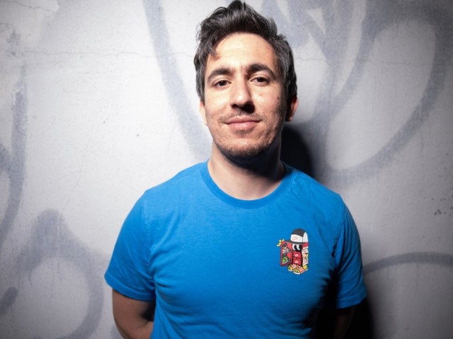 Smiling man in blue t-shirt with crest, standing against a gray graffiti wall