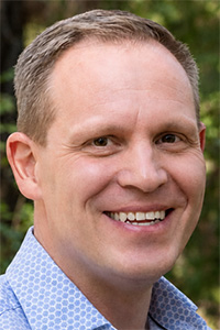 Smiling man with short blond hair, blue shirt, green outdoor background.