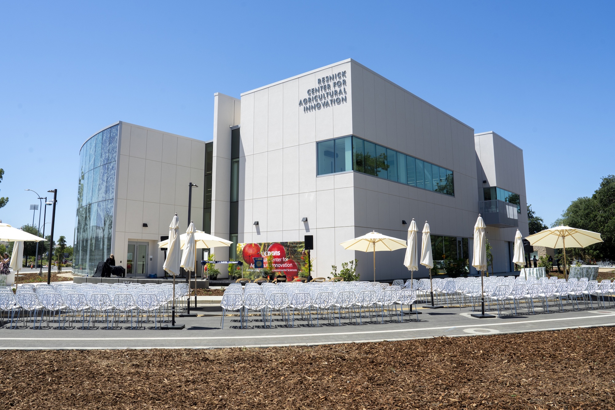 Modern white building photographed with outdoor seating, umbrellas, and clear blue sky. Exterior shot of the new Resnick Center for Agricultural Innovation at UC Davis. (TJ Ushing / UC Davis)