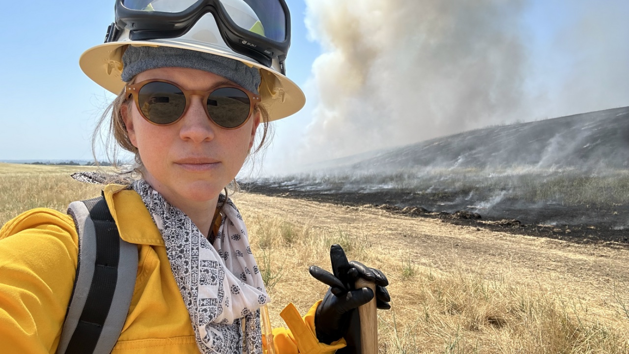 Emily Schlickman of UC Davis in yellow firefighting jumpsuiit, white hardhat, sunglasses looking at camera as smoke from prescribed burn billows from yellow field behind her