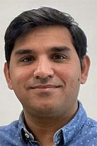 Headshot of smiling man with dark hair in blue collared shirt, light neutral background
