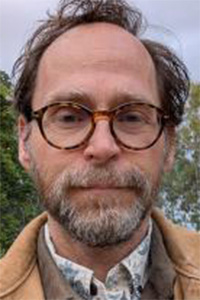 Portrait of man with round glasses, beard, and brown jacket outdoors
