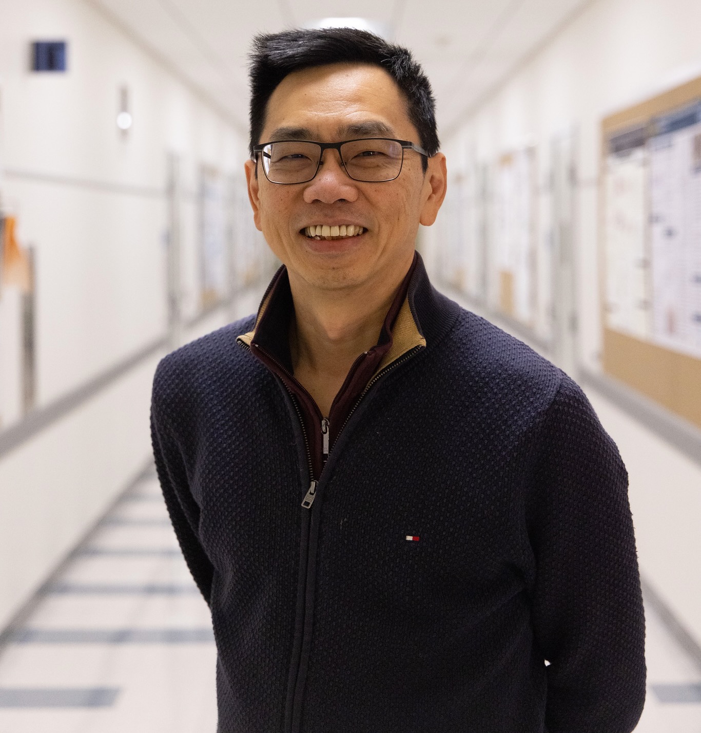 Scientist Cheemeng Tan wearing glasses and a dark zip sweater standing in a bright hallway