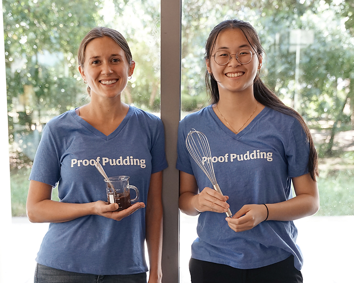 Two smiling women in matching 'Proof Pudding' tees holding a glass jar and whisk outdoors