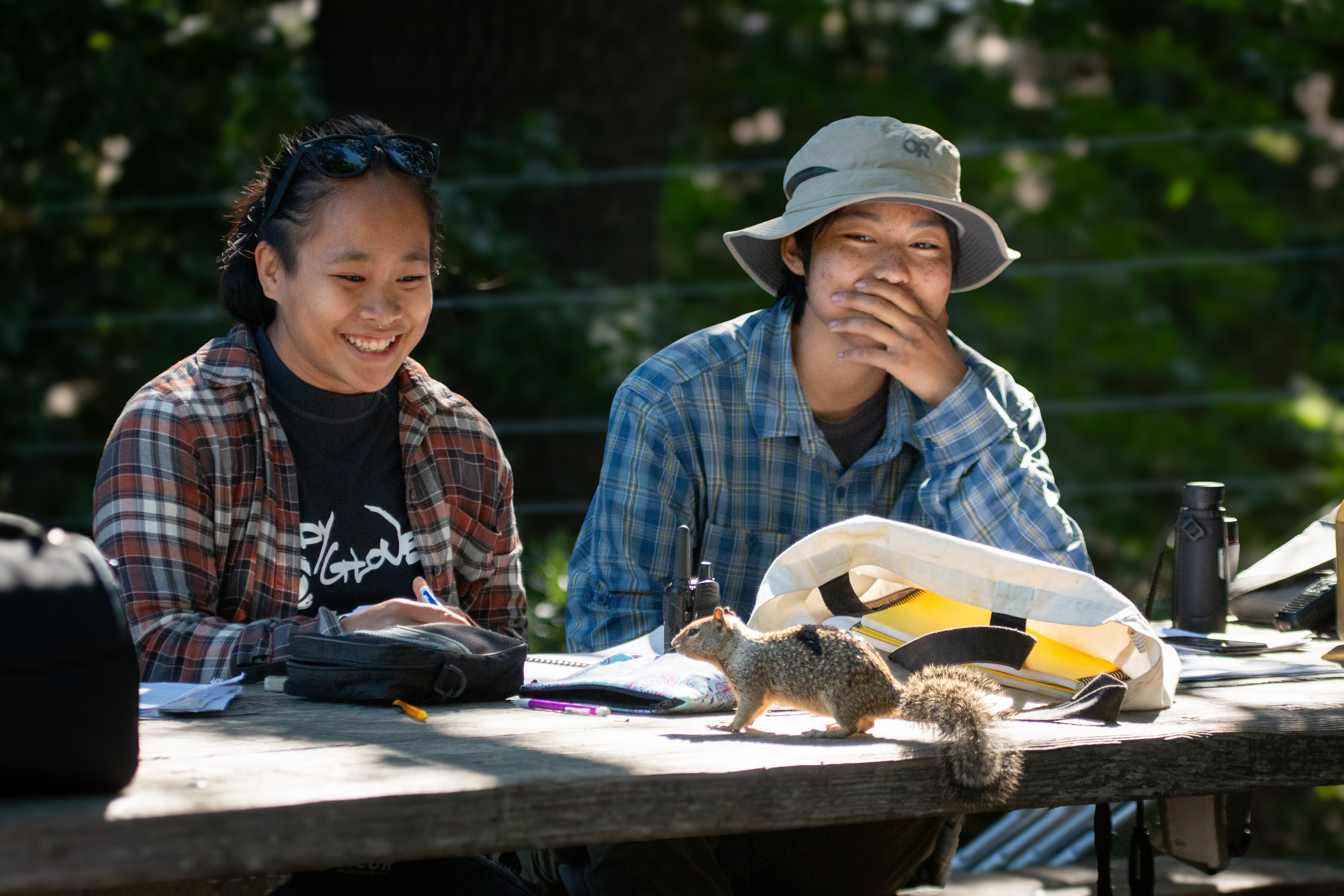 Tia Ravara in flannel shirt and black tee site beside Ryann Su in hat smiling as they watch a squirrel on their picnic table