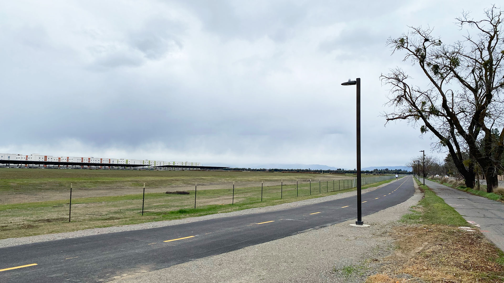 Photo of a bike path with a sidewalk on one side of it and fields on another. A distant development of apartments with solar panel covered parking can be seen.