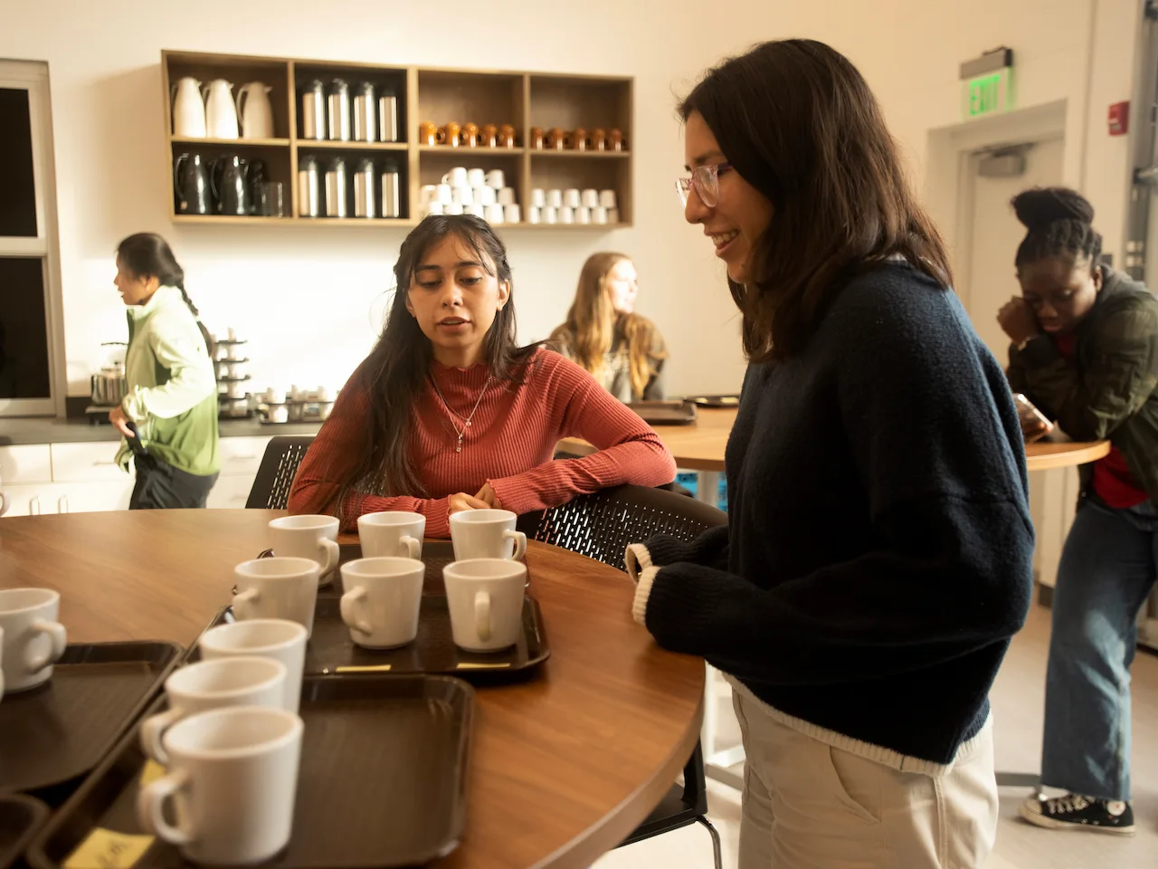 Two students talk at a wooden table with trays of white coffee mugs while other students mingle and check their phones behind them.