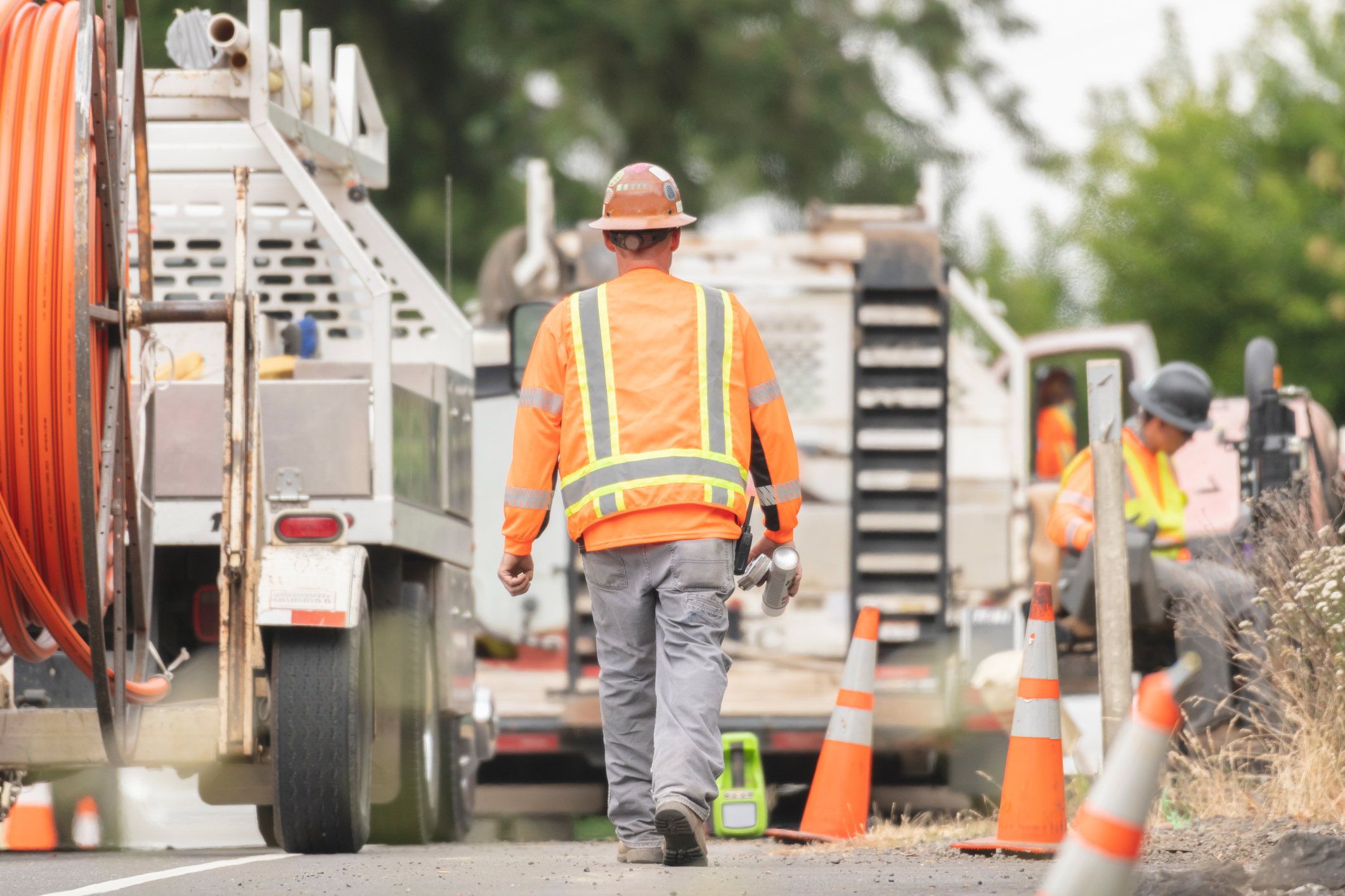Utility worker in orange vest and hard hat walking past trucks and traffic cones