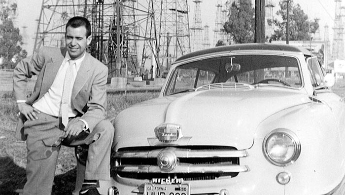 Old black and white photo of a man dressed in a suit with one foot up on the front bumper of a car.