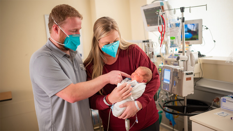 Masked parents in a hospital holding a swaddled newborn beside medical monitors
