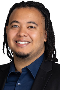 Smiling man with dreadlocks, wearing a black blazer and blue shirt against a white background.