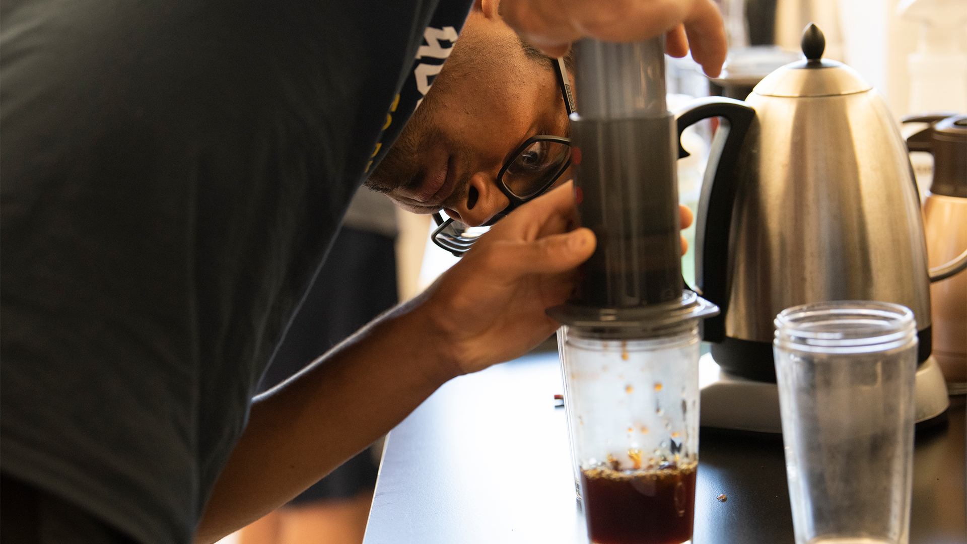 A student wearing a dark T-shirt and glasses leans over a lab table, which is filled with coffee and science instruments, to push hard on a device that drips coffee into a clear cup.