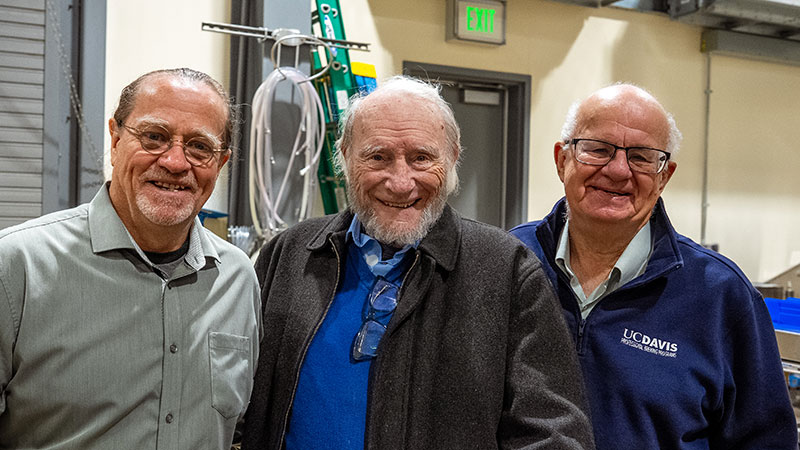 Three smiling men stand together, indoors, with tools and a ladder visible in the background.