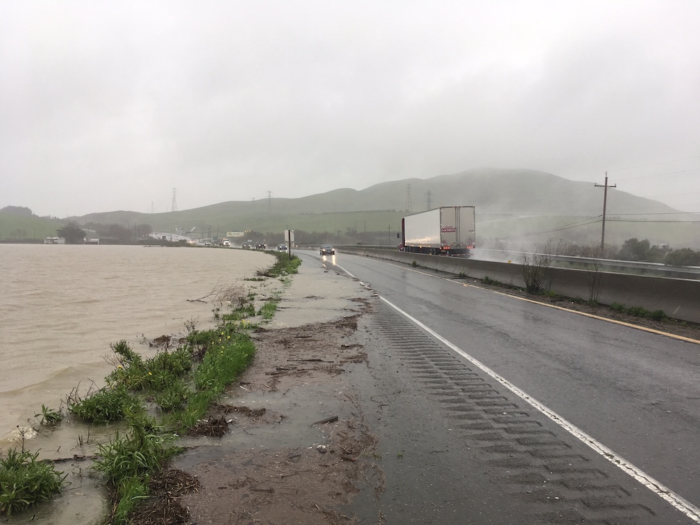 Flooded highway beside muddy river, trucks on wet lane under foggy green hills