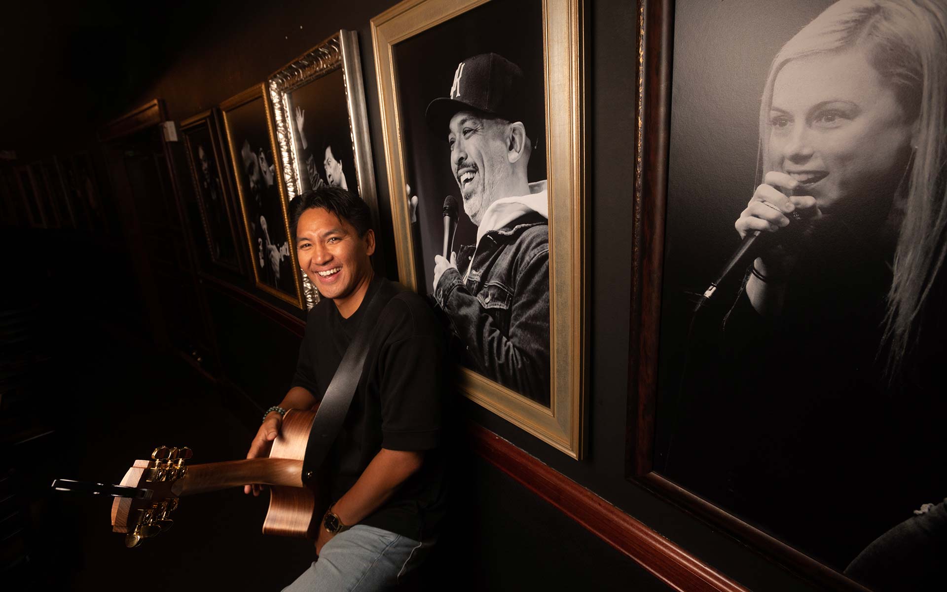 Photo of a man sitting and holding a guitar. He looks at the camera and smiles. On the wall behind him are framed black and white photos of comedians performing.