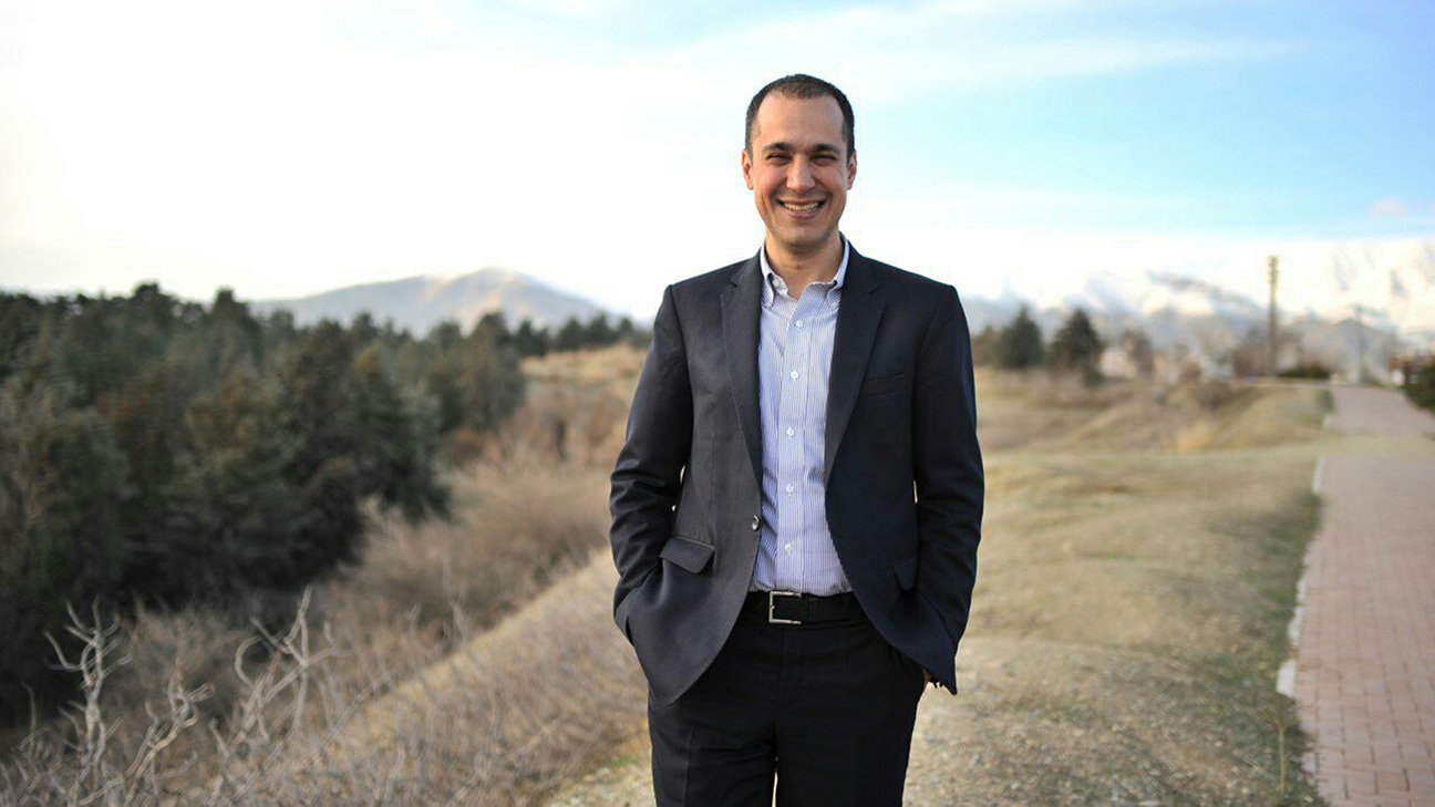 Smiling man in dark suit on a trail with trees and distant mountains, photo