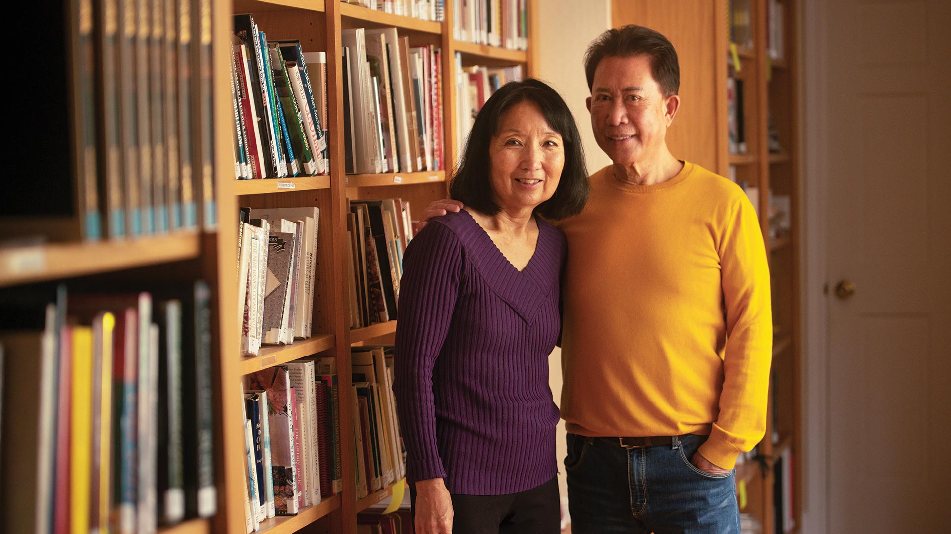 A woman and man stand in front of a large bookcase.